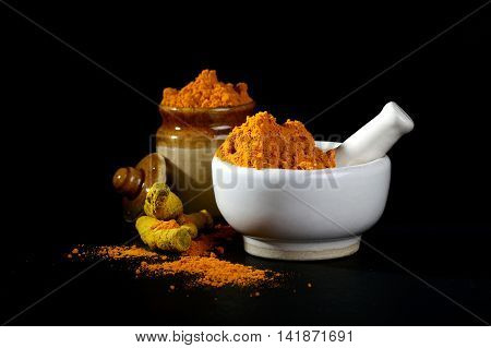 Turmeric powder in mortar with pestle and roots with clay pot on black background