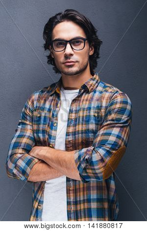 Confident young man. Confident young man keeping arms crossed and looking at camera while standing against grey background