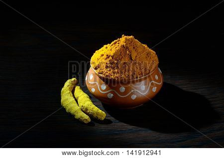 Turmeric powder in clay pot with roots or barks on black background