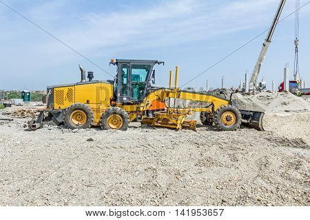 Grader is parked on gravel on construction site. Landscape transform into urban area with machinery people are working.