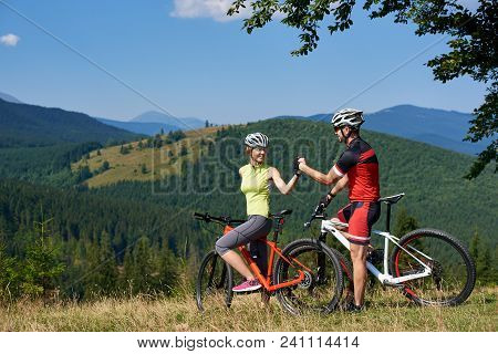 Happy Couple Cyclists, Man And Woman With Bicycles, Standing On Grassy Hill Under Big Green Tree Bra