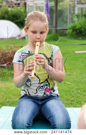 Portrait Of Beautiful Little Girl With Blond Hair In Jeans Playing Flute In The Garden Sitting On Th