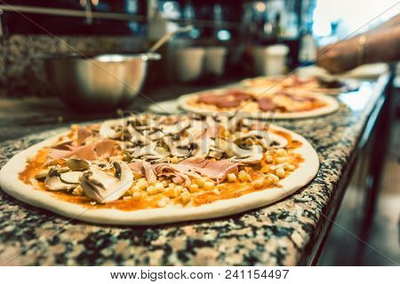Close-up of unbaked pizza dough topped with mushrooms, corn, ketchup and prosciutto on the counter in the kitchen of an Italian restaurant
