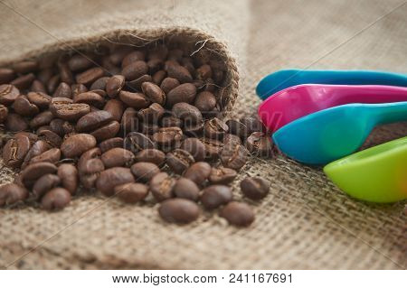 Loose Roasted Coffee Beans And Color Plastic Dose Measuring Spoons On Jute Sack. Conceptual Of Recom