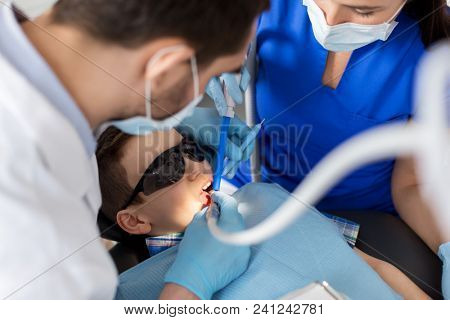 medicine, dentistry and healthcare concept - dentist and assistant with dental drill and saliva ejector treating kid patient teeth at dental clinic