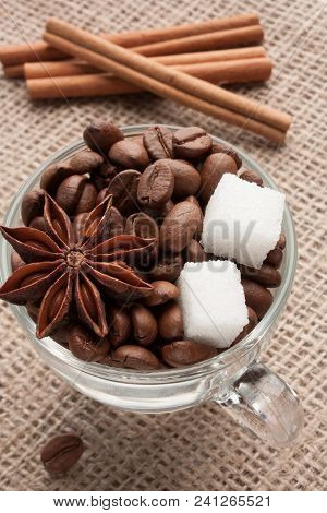Coffee Beans In Glass Cup With Sugar And Anise Star On Sackcloth With Cinnamon Sticks Behind. Fallen