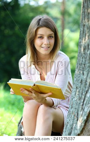 Happy blonde girl sitting and reading a book in summer park