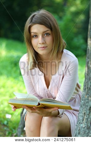 Happy blonde girl sitting and reading a book in summer park