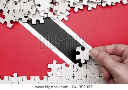 Trinidad And Tobago Flag  Is Depicted On A Table On Which The Human Hand Folds A Puzzle Of White Col