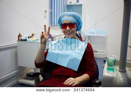 Smiling woman sits showing victory sign on dentist chair in clinic.