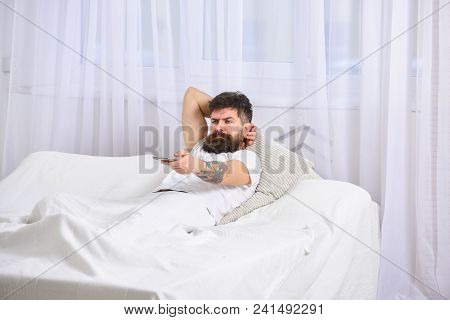 Man Laying On Bed, Watching Tv, White Curtains On Background. Guy On Serious Face Using Remote Contr