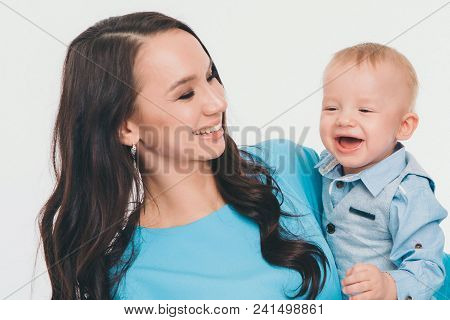 A Woman In A Blue Dress Holding A Baby In A Blue Shirt Against A White Wall. The Mother Took His Son