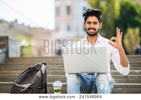 Happy Indian Student Sitting On The Stairs Showing Okay Sign Working On Laptop, In The University Ca