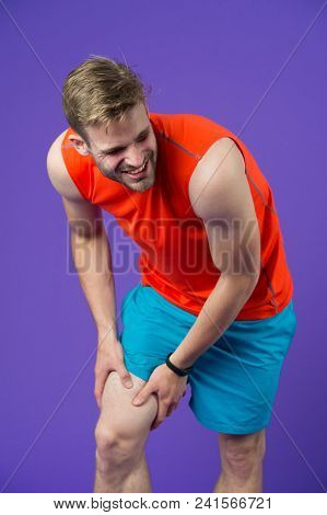 Sportsman With Knee Pain On Violet Background. Man In Orange Vest And Blue Shorts. Health Problem An