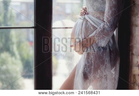 Pregnant Woman In A Beautiful White Peignoir And Lingerie. A Pregnant Woman Stands By The Window.