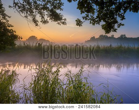 River The Dinkel With Lush Vegetation On Bank In Twente On An Early Summer Morning With Haze Over Th