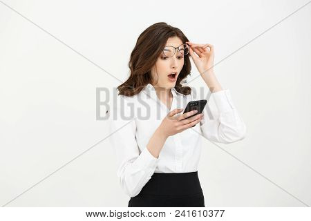 Business Concept: Surprised Youngwoman Holding Mobile Phone And Staring At It While Standing Against