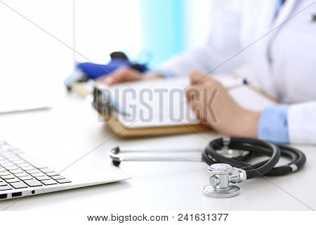 Closeup Of Stethoscope. Female Doctor Fills Up Medical Form While Sitting At The Desk In Hospital. H