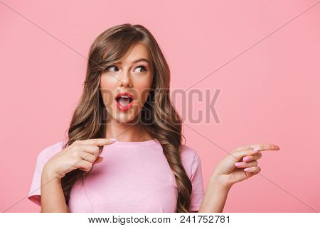 Closeup image of amusing brunette woman in basic clothing pointing finger aside at copyspace and presenting text or product isolated over pink background