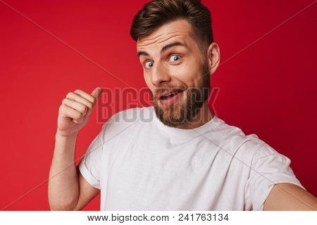 Picture of emotional young man standing isolated over red wall background make a selfie looking camera pointing to copyspace.