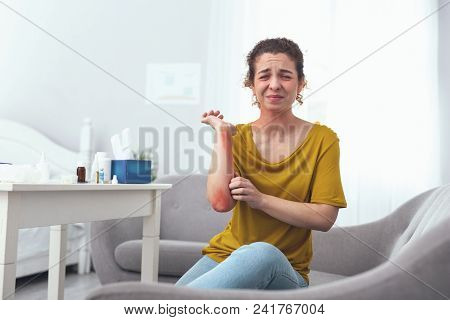 Itchy Elbow. Young Infected Girl Sitting On A Grey Couch Scratching Her Rashed Elbow Suffering From 