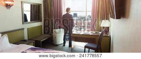 Young Businessman With Luggage In Hotel Room. Successful Man In Suit Looking At Window, Arriving To 