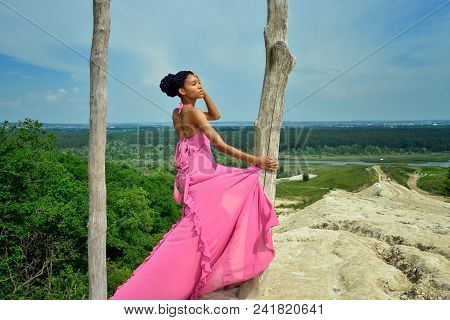 A Beautiful African American Girl, With Dreadlocks, Stands In A Pink Dress On A High Hill Against A 