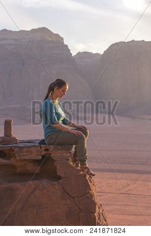 The Girl-traveler Sits On A Stone Ledge Against The Backdrop Of The Mountains In The Wadi Wadi Deser