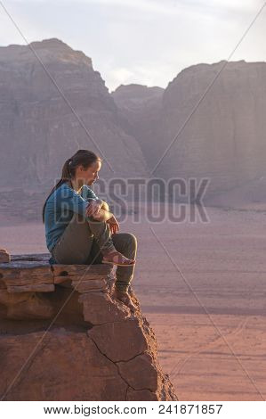 The Girl-traveler Sits On A Stone Ledge Against The Backdrop Of The Mountains In The Wadi Wadi Deser
