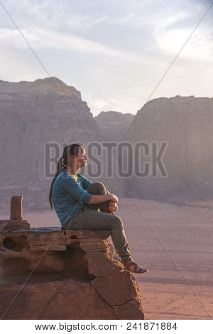 The Girl-traveler Sits On A Stone Ledge Against The Backdrop Of The Mountains In The Wadi Wadi Deser