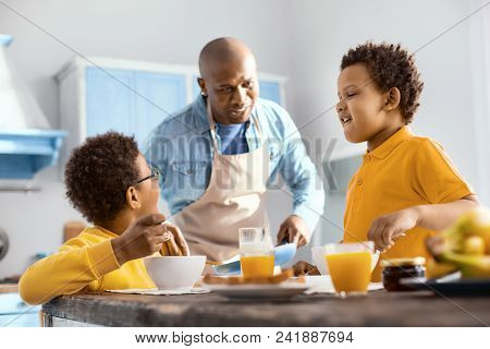 Mood-boosting Chat. Adorable Little Boys Chatting With Their Father And Laughing While Eating Cereal