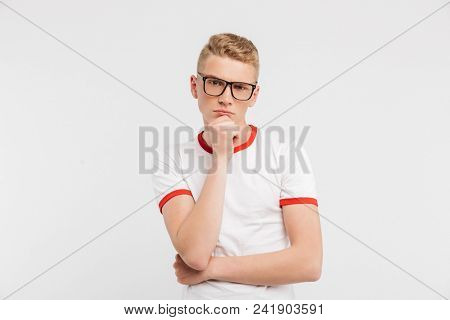 Image of smart serious guy wearing eyeglasses looking at camera with brooding gaze and touching chin, isolated over white background