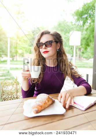 Beautifl Attractive Young Blonde Woman With Curly Hair Drinking Cappuccino And Eating Croissant In C