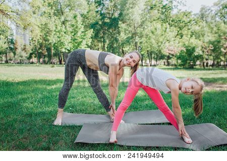 Cheerful And Beautiful Girls Are Excercising Outisde. They Are Curving To The Left Leg With Both Han