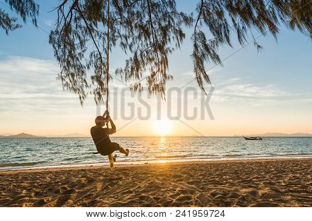 Man Sitting On A Swing At Beach During The Sunrise With A Beautiful Longtail Boat On The Sea.