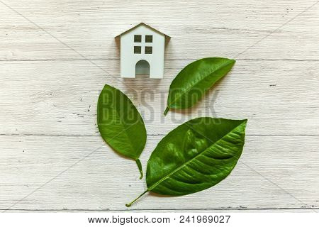 Miniature White Toy Model House With Green Leaves On Wooden Backgdrop. Eco Village, Abstract Environ