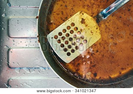 Dirty Greasy Steel Pan With Perforated Spatula. Placed On Top Of A Steel Kitchen Sink With Water Dro