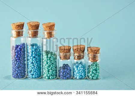 Purple And Cyan Beads In Glass Jars On Bright Blue Background. Beads In A Transparent Container With