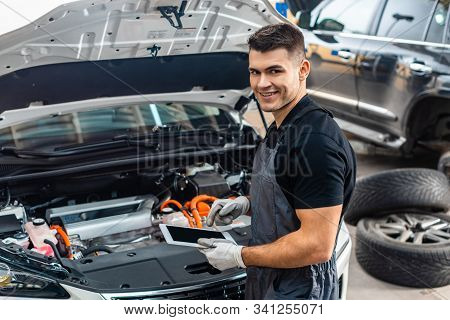 Handsome Mechanic Smiling At Camera While Using Digital Tablet Near Car Engine Compartment