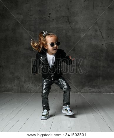 Cheerful Frolic Kid Girl In Leather Jacket, Stylish Jeans And Sunglasses Is Having Fun Playing Jumpi