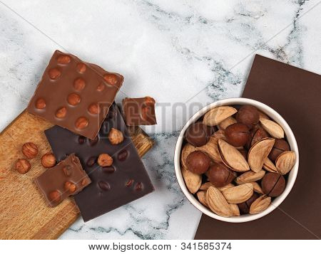 Broken Chocolate Bars, Hazelnuts And Raw Inshell Macadamia And Almonds On Gray Marble Background. To