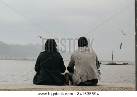 A Couple Sitting With Their Backs Ralaxing In Front Of The Sea With Mountaions And Seagulls In The B