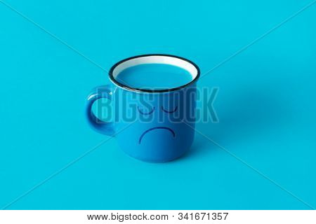 closeup of a blue mug, with a sad face drawn in it, and full of a blue liquid, on a blue background