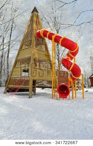 Big Toboggan With Wooden Pyramid Shaped Base And Red Twisted Tube On Snowy Playground In The Middle 