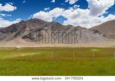 Vibrant Green Pasture With Yurt At The Foot Of Barren Rocky Mountains Under Cloudy Blue Sky In Pamir