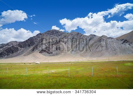 Vibrant Green Pasture With Yurt At The Foot Of Barren Rocky Mountains Under Cloudy Blue Sky In Pamir