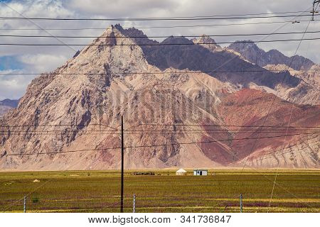 Vibrant Green Pasture And Flowers With Yurt At The Foot Of Barren Rocky Mountains Under Cloudy Sky I