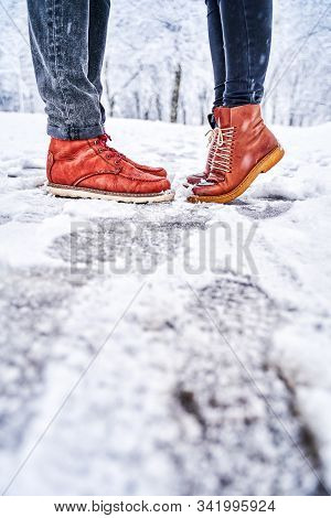 Feet Of A Couple On A Snowy Sidewalk In Brown Boots. Girl Stands On Toes While Kissing. Winter Weath
