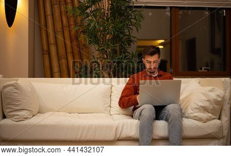 A Young Man With A Beard In A Red Shirt Works With A Laptop And Sits On The Couch In The Evening In 