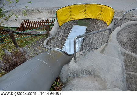 Stainless Steel Slide On The Field With Grassy Hills And Wooden Palisades. Large Glossy Tube For Sli